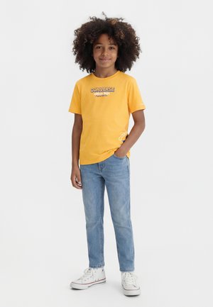 Jeune personne aux cheveux bouclés portant un t-shirt jaune Converse, un jean bleu clair et des baskets blanches montantes, debout avec une main dans la poche.