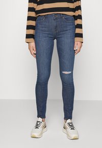 Calças de ganga slim fit azuis com um detalhe rasgado nos joelhos, combinadas com ténis creme e pretos. O tecido tem uma textura suave e ligeira elasticidade.