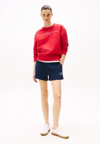 Rode sweatshirt met witte tekst en marineblauwe shorts, beide in zachte materialen. Casual sneakers in het wit met tan-kleurige zolen complementeren de outfit.