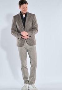 Brauner Hahnentrittmuster-Blazer mit strukturierter Oberfläche, kombiniert mit hellgrauen Hosen und weißen Turnschuhen. Neutrale Farbpalette.