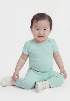 Småbarn med kort, mørkt hår som sitter med beina krysset, iført en mintgrønn ribbestrikket dress og hvite joggesko, smilende mot kameraet.