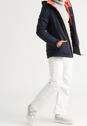 Veste matelassée bleu marine avec accents corail et capuche, associée à un pantalon blanc et des bottes isolantes gris clair, présentant un design élégant.