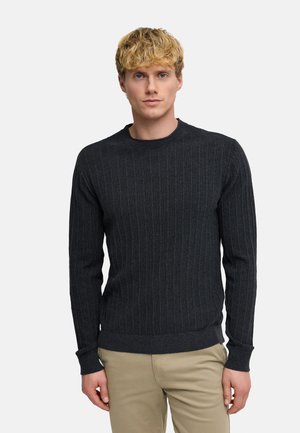 Blondyn w ciemnoszarym prążkowanym swetrze i beżowych spodniach, stojący na tle jednolitego jasnoszarego tła.