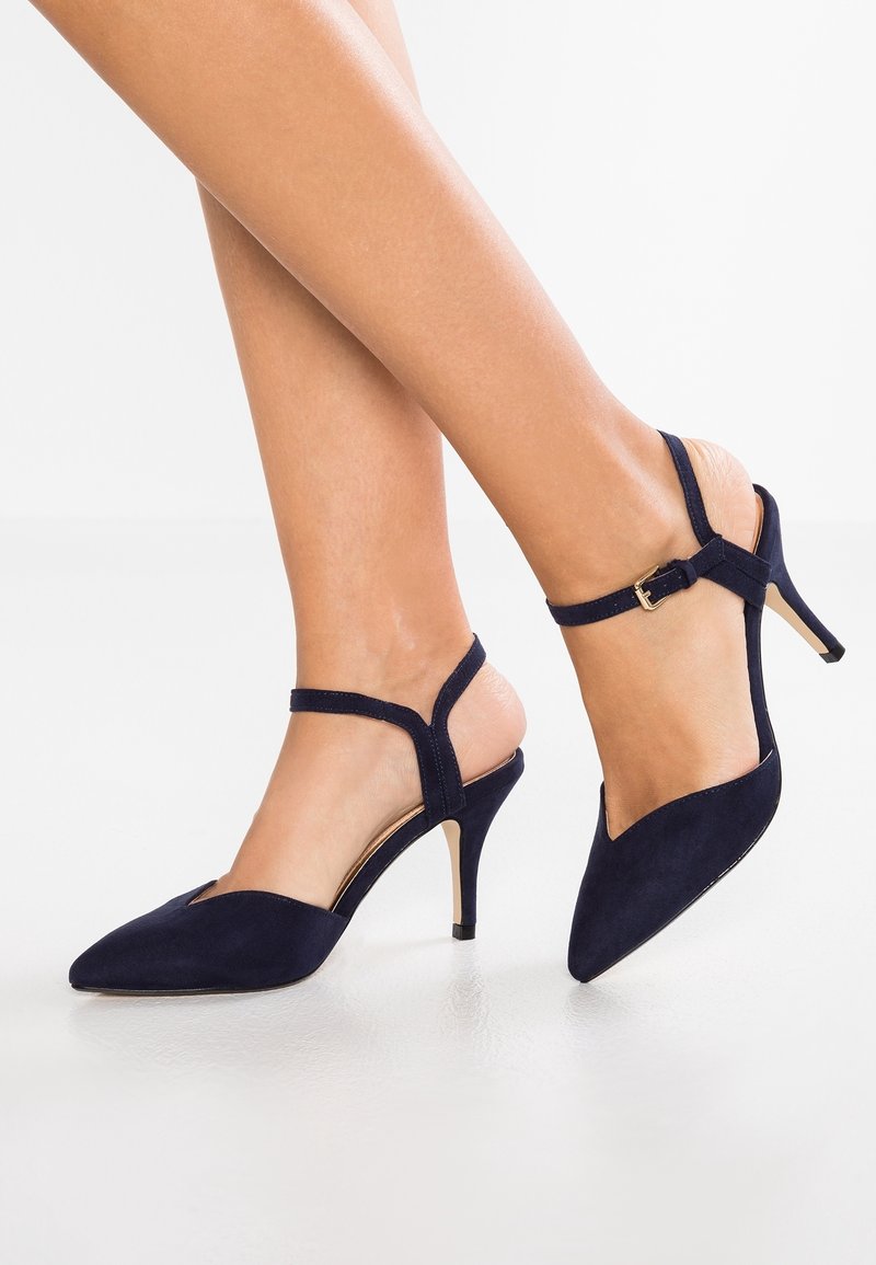 Jambes portant des escarpins noirs à bout pointu avec des brides à la cheville et de petites boucles dorées, debout sur un sol blanc.