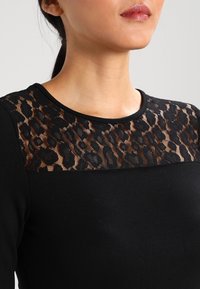 Maglietta nera a maniche lunghe con un pannello in pizzo trasparente leopardato sul collo, caratterizzata da una texture liscia e un design aderente.