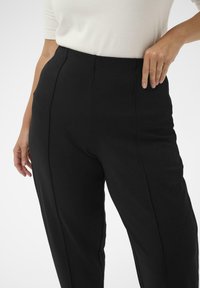 Pantaloni negri cu un design drept, țesătură texturată și pliuri frontale. Mâna modelului este așezată pe talie.
