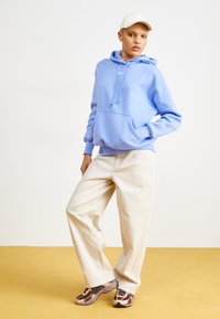 Osoba u svijetloplavoj dukserici, kremastim hlačama širokih nogavica, smeškim tenisicama i bijelom šeširu, stoji na žutoj podlozi nasuprot bijelom zidu.