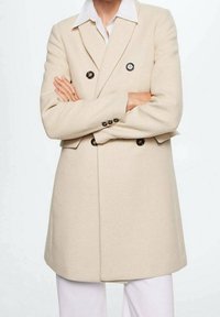 Cappotto beige doppiopetto realizzato in tessuto morbido, caratterizzato da grandi bottoni neri, una vestibilità sartoriale e una silhouette strutturata. Camicia bianca sotto.