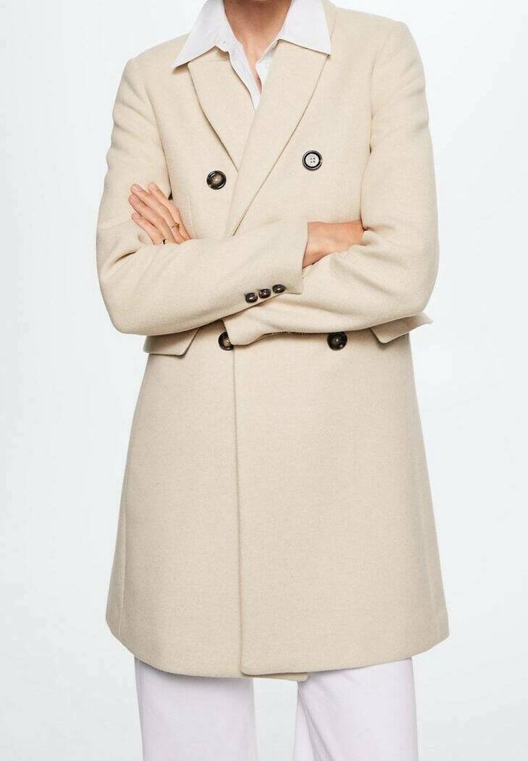Cappotto beige doppiopetto realizzato in tessuto morbido, caratterizzato da grandi bottoni neri, una vestibilità sartoriale e una silhouette strutturata. Camicia bianca sotto.