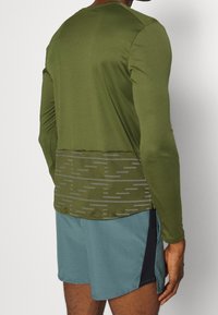 Mann trägt ein olivgrünes Langarmshirt mit reflektierenden Streifen im unteren Rückenbereich und hellblaue Sportshorts mit schwarzen Seitenpanelen.