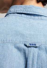 Lichtblauw denim shirt met een puntkraag, een achterplooi en een klein blauw label bij de halslijn. Zachte, textuurrijke stof.