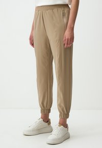 Beige joggerbroeken met een elastische tailleband, zijzak met rits en taps toelopende boorden. Gemaakt van lichtgewicht materiaal met een gladde textuur.