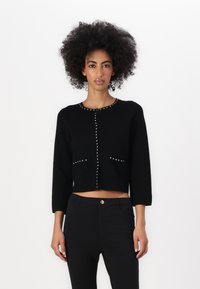 Svart cropped cardigan med trekvartslange ermer, prydet med sølvstifter langs fronten og lommene. Myk tekstur, tettsittende design.
