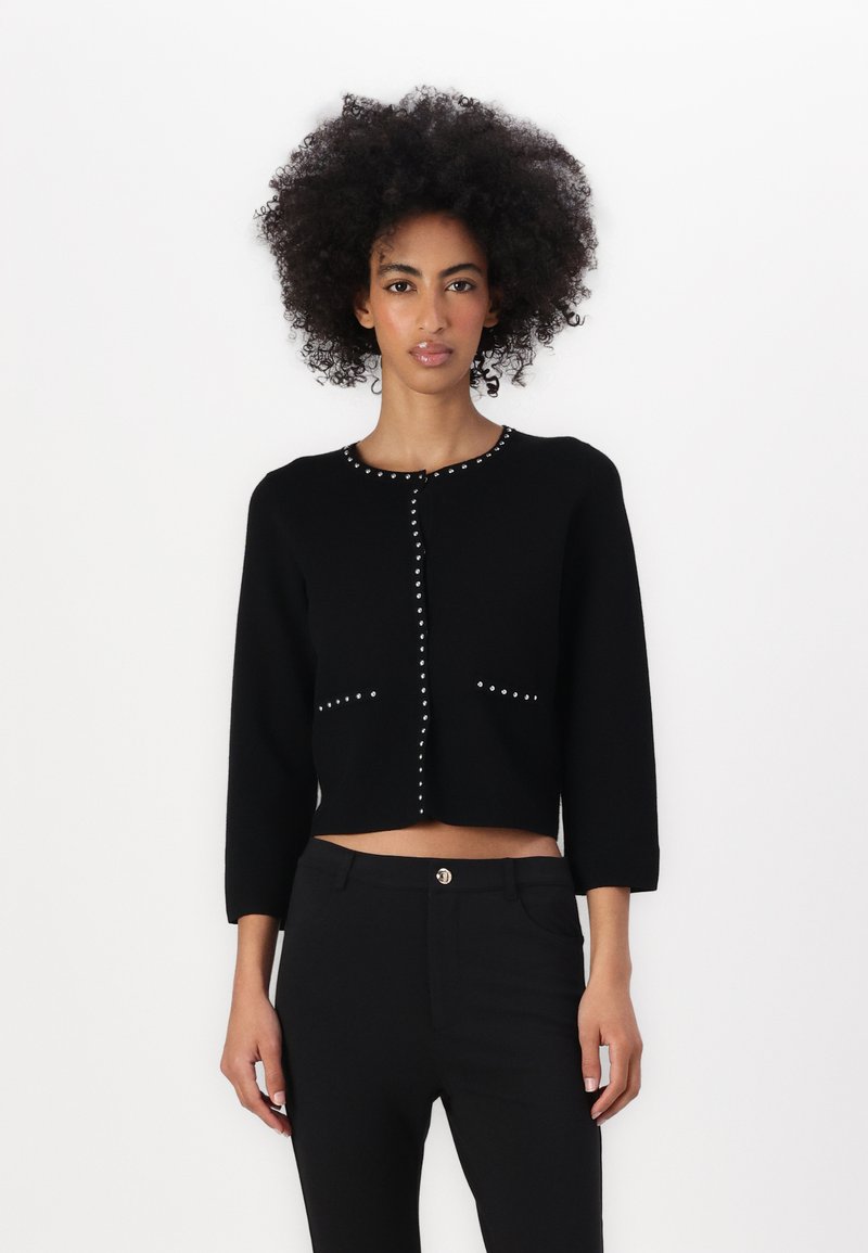 Svart cropped cardigan med trekvartslange ermer, prydet med sølvstifter langs fronten og lommene. Myk tekstur, tettsittende design.