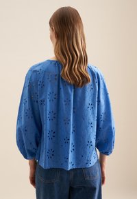 Blå bomuldsbluse med pufærmer, med indviklede blomsterudskæringer og en afslappet pasform. Glat tekstur, afslappet design.