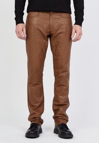 Braune Lederhosen mit glatter Textur, schmalem Beinansatz und dezenten Nähdetails. Getragen mit schwarzen Schuhen und einem schwarzen Pullover.