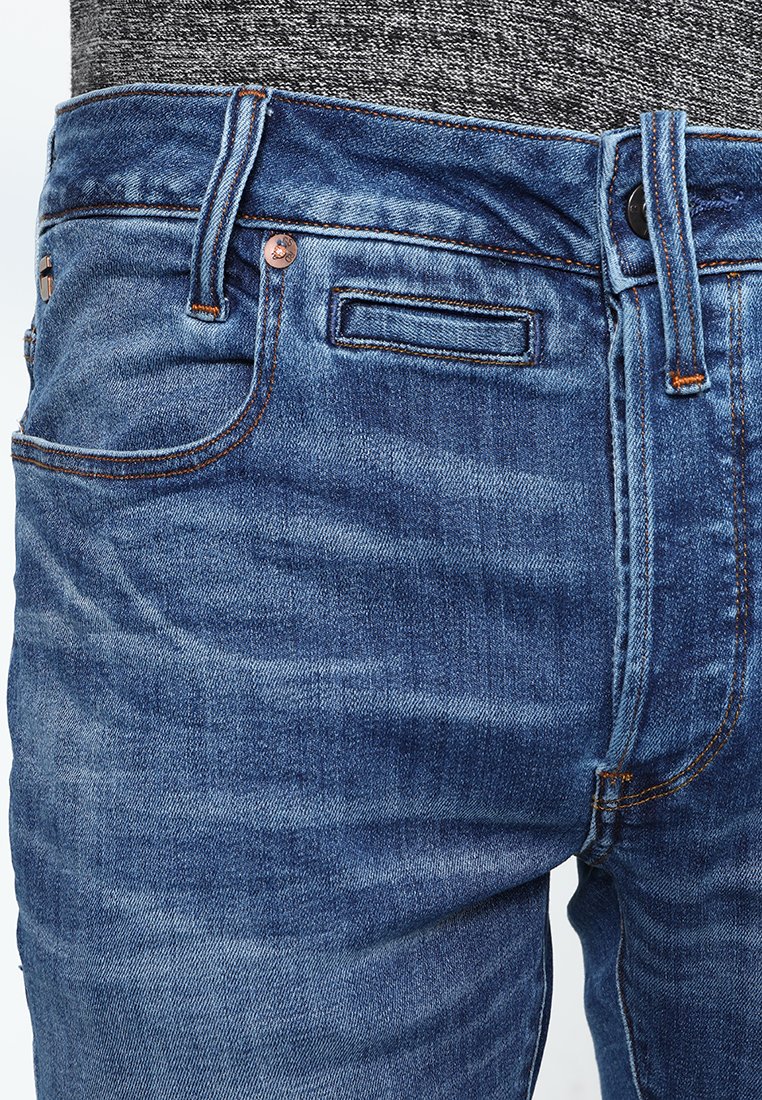 Jeans en denim bleu avec une coupe classique, dotés d'une fermeture à bouton, de passants de ceinture, de poches avant et de coutures orange contrastantes.