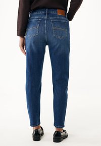 Žena oblečená v modrých džínech s vysokým pasem, které mají zadní kapsy a béžové švy. K džínsům má na sobě černé mokasíny a tmavý dlouhý rukáv top, stojí.