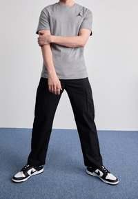 Šedé tričko s krátkým rukávem a černým logem, černé cargo kalhoty a černo-bílé tenisky na modré textilní podlaze.