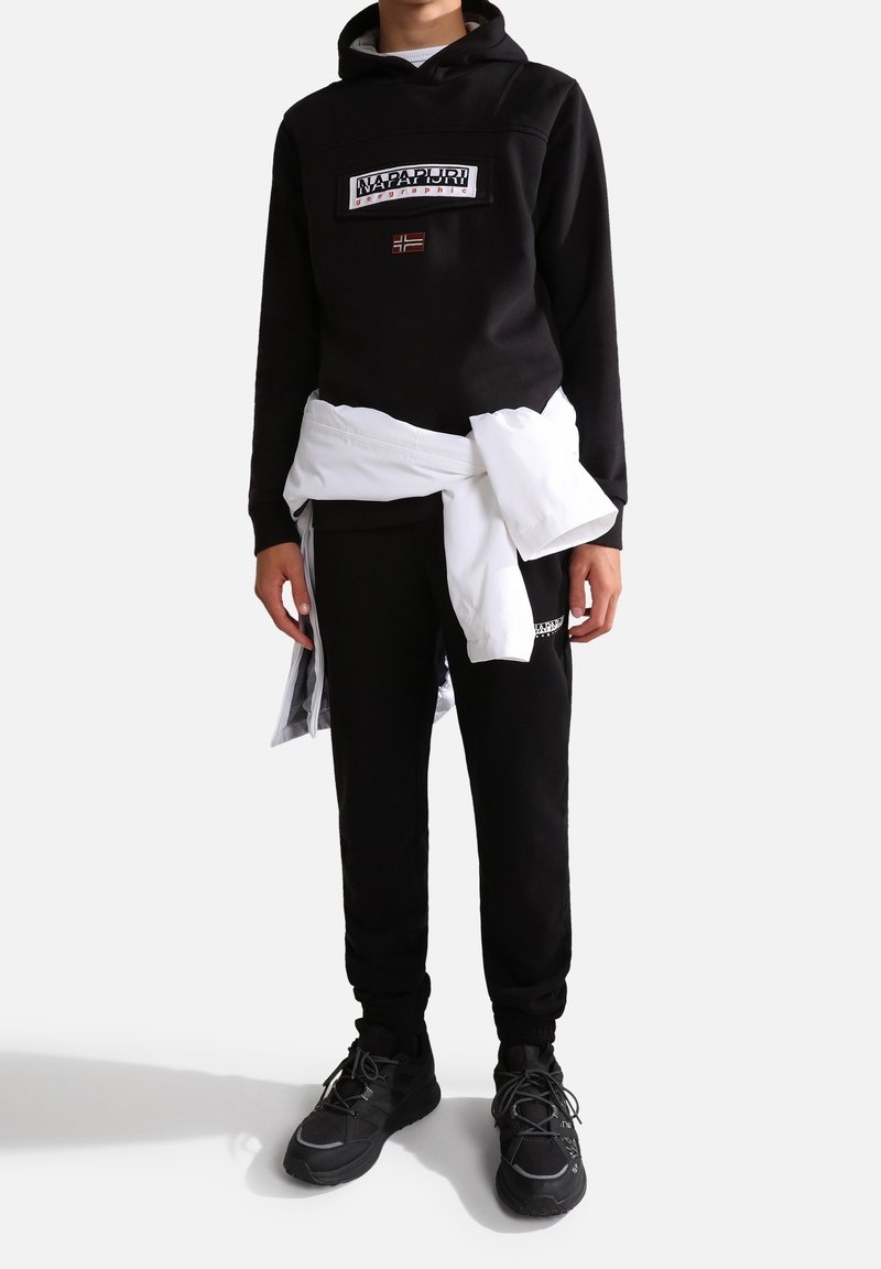 Personne portant un sweat à capuche et un pantalon noirs de la marque Napapijri avec une veste blanche nouée autour de la taille, debout devant un fond uni.