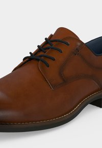 Chaussure habillée en cuir marron avec lacets noirs, coutures visibles et semelle noire sur fond clair.