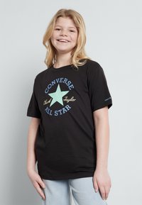 Camiseta de algodón negra con un gráfico colorido que presenta una estrella verde claro y el texto "Converse Chuck Taylor All Star". Mangas cortas y ajuste relajado.