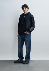 Vīrietis stāv, valkājot melnu sporta džemperi ar kapuci ar rakstiem uz piedurknēm, zilas labotas džinsas ar burtiem un melnas sporta kurpes pret vienkāršu fonu.