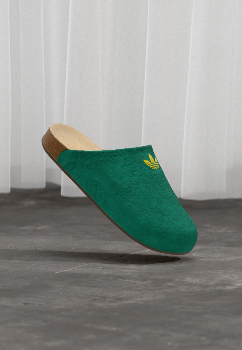 Zöld velúr bebújós cipő virágos textúrával, fakóbarna talpbetéttel és barna gumitalppal. Sárga logó díszíti a felsőrész oldalát.