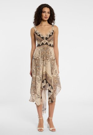 Vrouw met krullend haar, gekleed in een beige jurk met patroon en een sjaalzoom, en beige sandalen met hoge hakken, staand tegen een witte achtergrond.