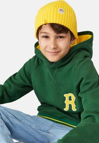 Grön bomullshuvtröja med gula accenter och en "R"-logotyp, bärs tillsammans med en ribbad gul mössa. Blå denimjeans syns också.