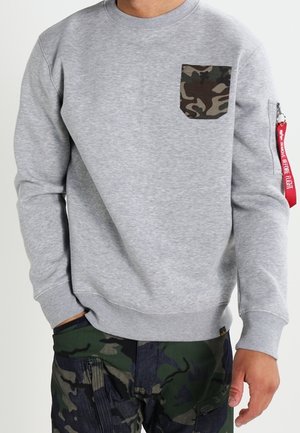 Mand iført grå sweatshirt med camouflage-lomme og camouflage cargobukser, stående med den ene hånd ned langs siden mod ensfarvet baggrund.
