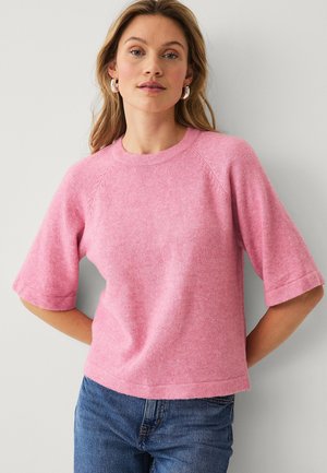 Vrouw die een roze trui met korte mouwen en een blauwe spijkerbroek draagt, staand tegen een lichtgrijze muur met haar handen achter haar rug.