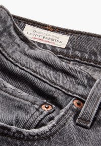 Jeans de mezclilla gris con una superficie texturizada, que presentan costuras de color óxido, remaches de cobre y una etiqueta que dice "Levi's Premium, San Francisco, CA."