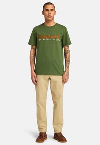 Zelené bavlněné tričko s logem "Timberland" v oranžové barvě, spárované s khaki kalhotami a hnědými šněrovacími botami, stojící na neutrálním pozadí.