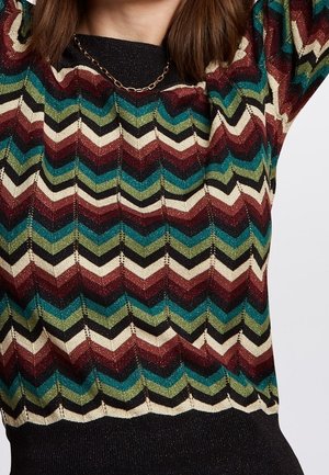 Gros plan sur un pull à rayures zigzag multicolores en rouge, vert, bleu, noir et blanc, porté par-dessus un haut noir avec un collier chaîne en or.
