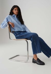 Lichtblauw gestreept overhemd met knopen; wijde blauwe spijkerbroek; zwarte loafers; zittend op een chromen en houten cantileverstoel tegen een lichte achtergrond.