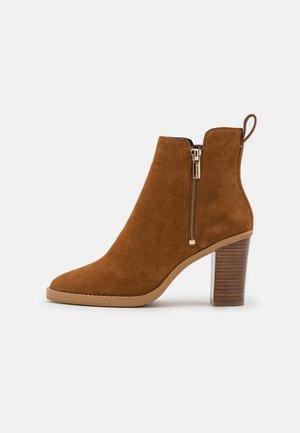 Brauner Ankle-Boot aus Wildleder mit Reißverschluss an der Seite, Schlaufe zum Anziehen, geschichtetem Blockabsatz und rundem Zehenbereich, im Profil vor weißem Hintergrund gezeigt.