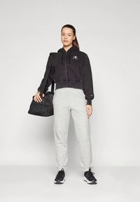 Mujer con sudadera negra con cremallera, pantalones de chándal grises y zapatillas negras, sosteniendo un bolso de deporte negro sobre su hombro, contra un fondo liso.