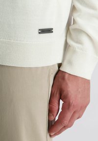 Helles cremefarbenes Sweatshirt mit gerippten Bündchen und Saum. Enthält ein kleines schwarzes Etikett mit weißem Text in der linken unteren Ecke. Getragen über beigen Hosen.