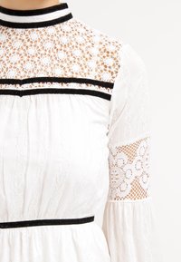 Biała sukienka z koronkowym dekoltem, czarnymi aksamitnymi paskami i koronkowymi akcentami na rękawach. Ma teksturowany materiał i wysoki dekolt.