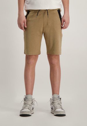 Persoon in beige drawstring shorts met zwarte ritszakken en witte high-top sneakers, staande op een effen achtergrond.