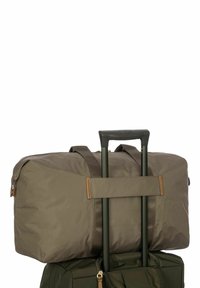 Sac de voyage vert olive avec une finition mate, équipé d'accents en cuir marron, de sangles réglables et monté sur un châssis à roulettes noir.