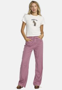 Witte cropped t-shirt met de tekst "KISS MY" en een cartoon-ezelafbeelding, gecombineerd met lichtroze wijde broek en bruine schoenen.