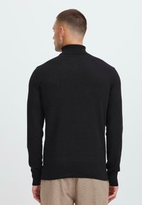 Hombre vestido con un suéter negro de cuello alto y pantalones beige, visto desde atrás sobre un fondo claro y liso.