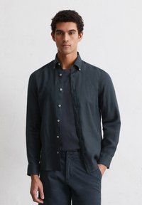 Donkerblauw, langemouwen shirt van linnen, met een button-down kraag en lichtgekleurde knopen, gedragen over een donker shirt.