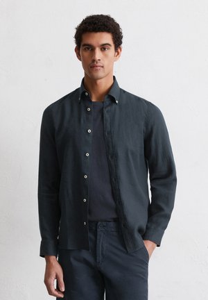 Donkerblauw, langemouwen shirt van linnen, met een button-down kraag en lichtgekleurde knopen, gedragen over een donker shirt.
