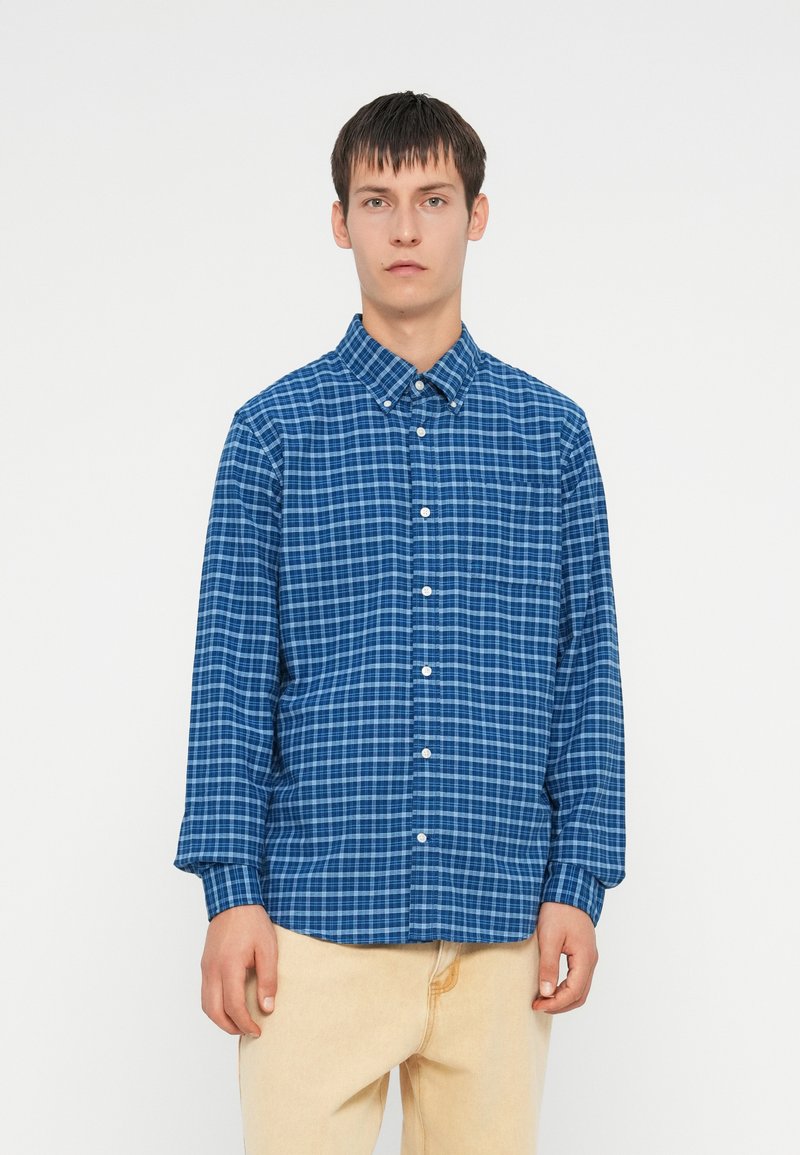 Camisa de cuadros azul de manga larga, fabricada con un tejido de patrón a cuadros. Cuello button-down y un bolsillo en el pecho.
