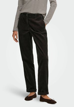 Femme portant un pantalon en velours côtelé noir, une chemise rayée à manches longues et des ballerines foncées, debout avec une main dans la poche.