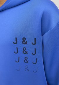 Modrá mikina s hladkým materiálem, na přední straně s opakovaným černým potiskem písmen "J & J" v různých velikostech.