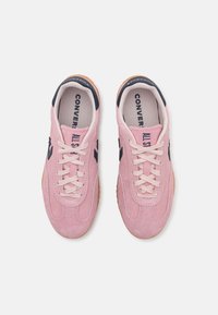 Para różowych zamszowych trampek Converse All Star z białymi sznurowadłami, czarnymi detalami logo i gumowymi podeszwami widziana z góry.